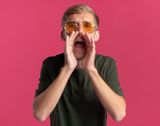 Young handsome guy looking at front wearing green shirt and glasses calling someone isolated on pink wall