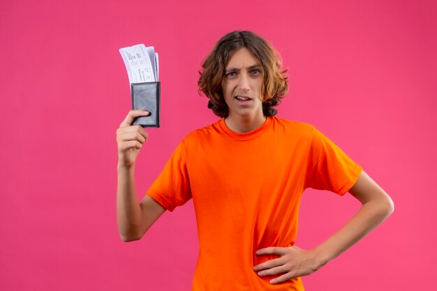 Young handsome guy in orange t-shirt holding air tickets holding air tickets displeased standing over pink background