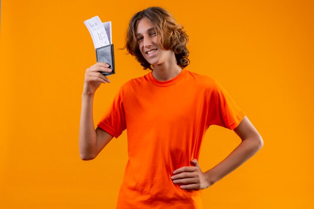 Young handsome guy in orange t-shirt holding air tickets looking at camera with confident smile standing