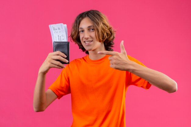Young handsome guy in orange t-shirt holding air tickets looking confident pointing with finger to it smiling cheerfully standing