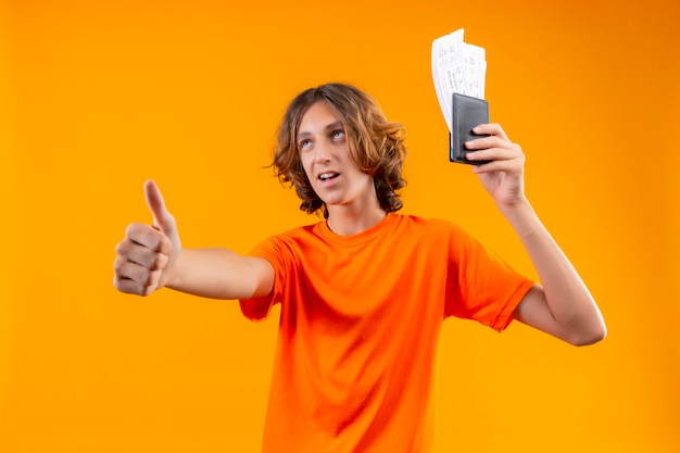 Молодой красивый парень в оранжевой футболке с билетами на самолет выглядит уверенно, показывает палец вверх, весело улыбаясь стоя