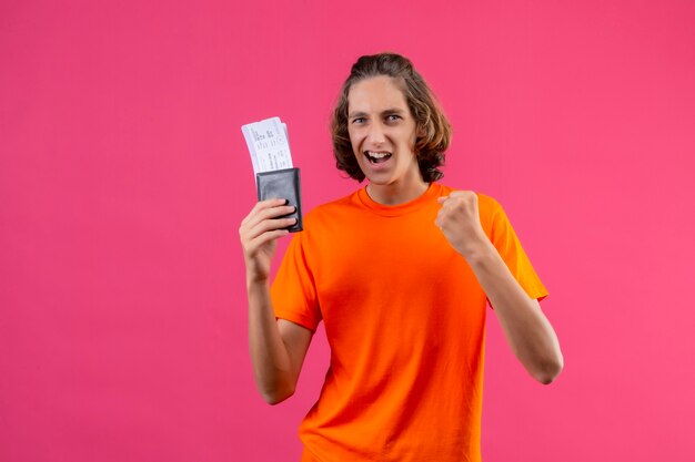 Young handsome guy in orange t-shirt holding air tickets raising fist smiling cheerfully winner concept standing over pink background