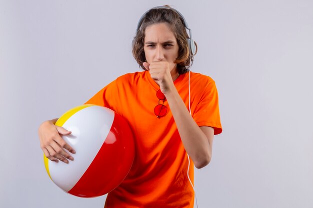 白い背景の上に立って体調不良の咳をしてヘッドフォンでインフレータブルボールを保持しているオレンジ色のtシャツの若いハンサムな男