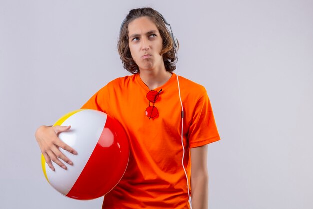 Молодой красивый парень в оранжевой футболке держит надувной мяч в наушниках, недовольно глядя в сторону с хмурым лицом, стоящим на белом фоне
