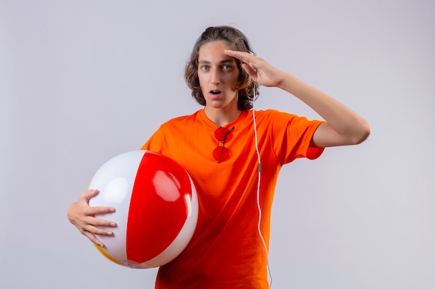 Free photo young handsome guy in orange t-shirt holding inflatable ball with headphones looking surprised standing with hand near head