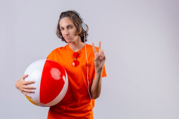 Young handsome guy in orange t-shirt holding inflatable ball with headphones showing number two smiling standing 