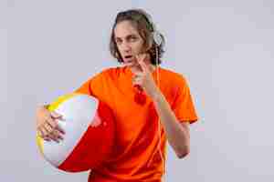 Free photo young handsome guy in orange t-shirt holding inflatable ball with headphones standing with pensive look looking uncertain over white background