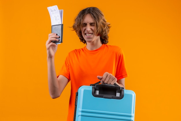 Young handsome guy in orange t-shirt holding travel suitcase and air tickets standing with closed eyes making desirable wish over yellow background