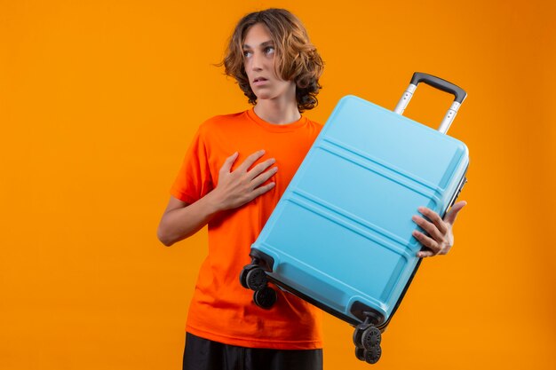 Young handsome guy in orange t-shirt holding travel suitcase looking aside with confused expression standing