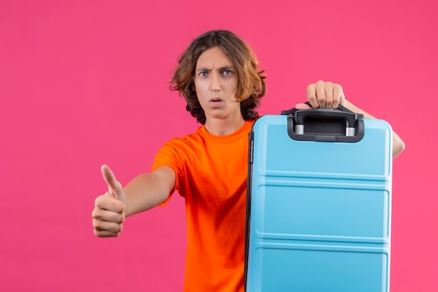 Young handsome guy in orange t-shirt holding travel suitcase looking at camera surprised showing thumbs up standing over pink background