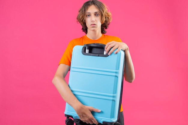 Young handsome guy in orange t-shirt holding travel suitcase looking at camera with sad face standing over pink background