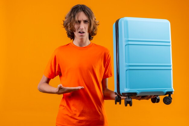 Young handsome guy in orange t-shirt holding travel suitcase pointing with arm of hand looking confused standing