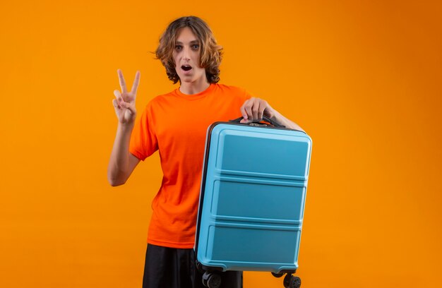 Young handsome guy in orange t-shirt holding travel suitcase showing number two or victory sign looking surprised standing over yellow background