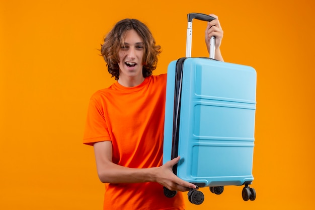 Young handsome guy in orange t-shirt holding travel suitcase smiling cheerfully positive and happy ready to travel standing