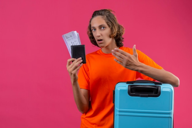 Young handsome guy in orange t-shirt standing with travel suitcase holding air tickets looking confused with arm raised over pink background