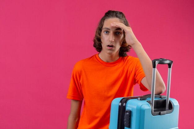 Free photo young handsome guy in orange t-shirt standing with travel suitcase looking confused and surprised with hand on head over pink background