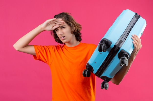 Free photo young handsome guy in orange t-shirt standing with travel suitcase looking tired and bored over pink background