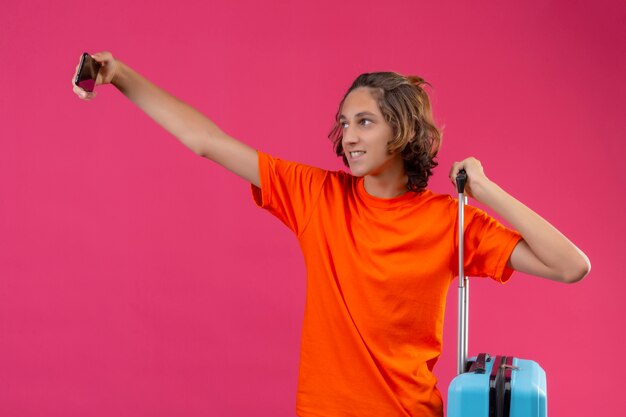 Young handsome guy in orange t-shirt standing with travel suitcase taking selfie using smartphone happy and positive