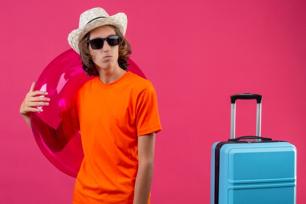 Young handsome guy in orange t-shirt and summer hat wearing black sunglasses holding inflatable ring looking aside with frowning face standing with travel suitcase