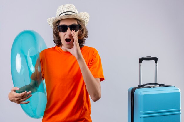 Young handsome guy in orange t-shirt wearing black sunglasses holding inflatable ring happy and positive shouting or calling someone with hand near mouth standing with travel suitcase over whit