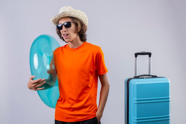 Young handsome guy in orange t-shirt wearing black sunglasses holding inflatable ring happy and positive sticking out tongue standing with travel suitcase 