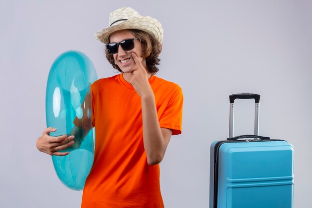 Young handsome guy in orange t-shirt wearing black sunglasses holding inflatable ring smiling cheerfully pointing with finger to his cheek standing with travel suitcase over white background