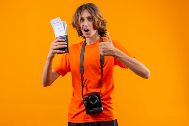 노란색 배경 위에 서있는 엄지 손가락을 유쾌하게 보여주는 항공 티켓을 들고 카메라와 함께 주황색 티셔츠에 젊은 잘 생긴 남자