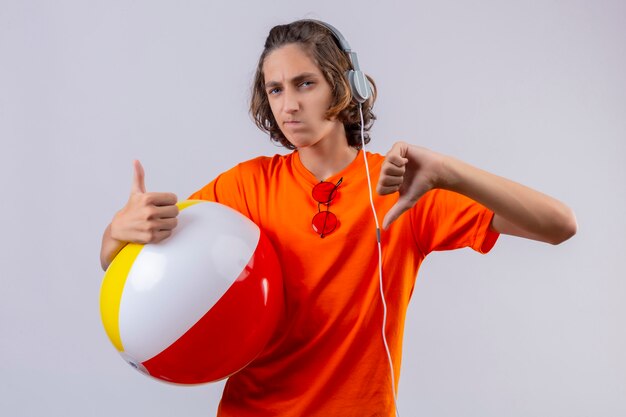 Young handsome guy in orange t-shirt with headphones holding inflatable ball displeased showing thumbs up and down standing 