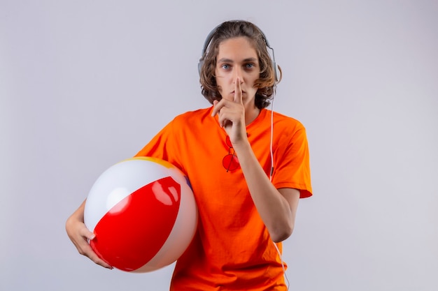 Young handsome guy in orange t-shirt with headphones holding inflatable ball making silence gesture with finger on lips standing over white background