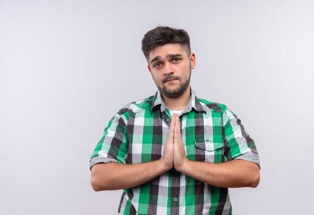 Young handsome guy wearing checkered shirt asking sincerely standing over white wall