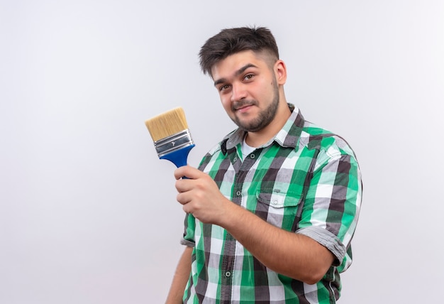 Young handsome guy wearing checkered shirt confidently looking holding paint brush standing over white wall