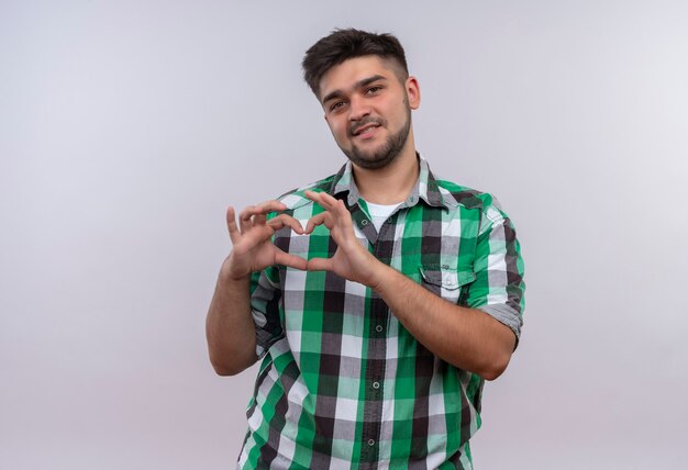 Young handsome guy wearing checkered shirt doing love sign with hands standing over white wall