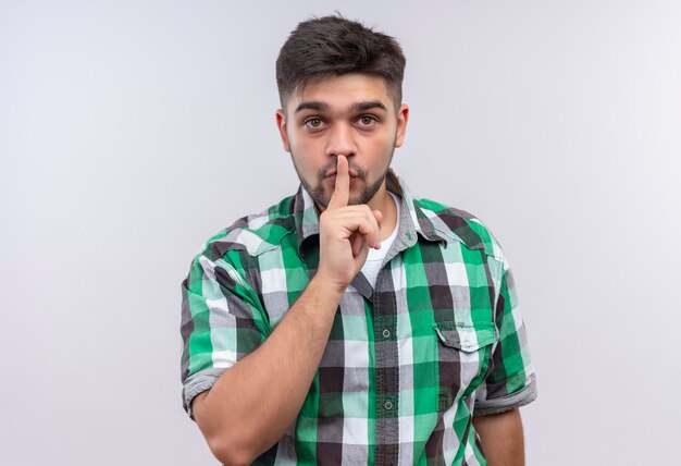 Young handsome guy wearing checkered shirt doing silence sign standing over white wall