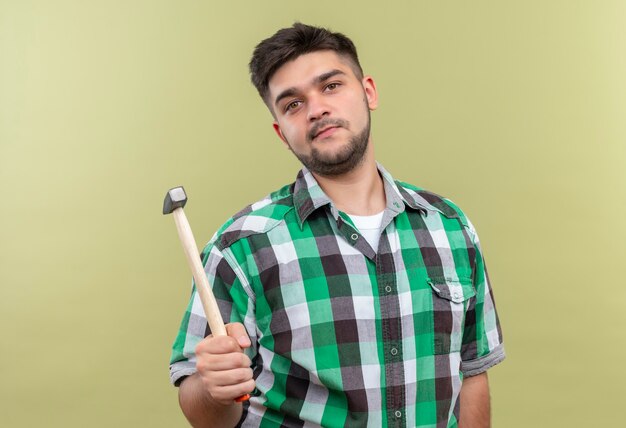 Young handsome guy wearing checkered shirt holding hammer standing over khaki wall