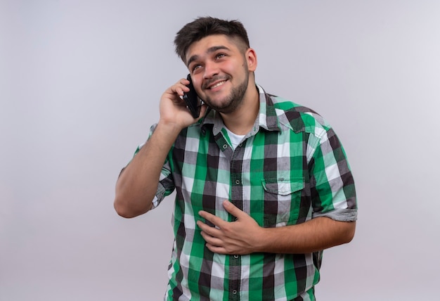 Young handsome guy wearing checkered shirt laughing and talking on phone standing over white wall