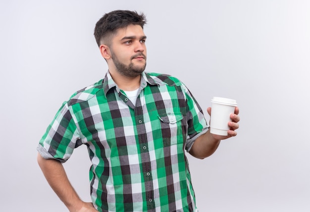 Молодой красивый парень в клетчатой рубашке смотрит, кроме того, держит пластиковую кофейную чашку, стоящую над белой стеной