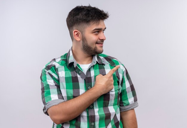 Young handsome guy wearing checkered shirt looking besides pointing to the left with forefinger standing over white wall