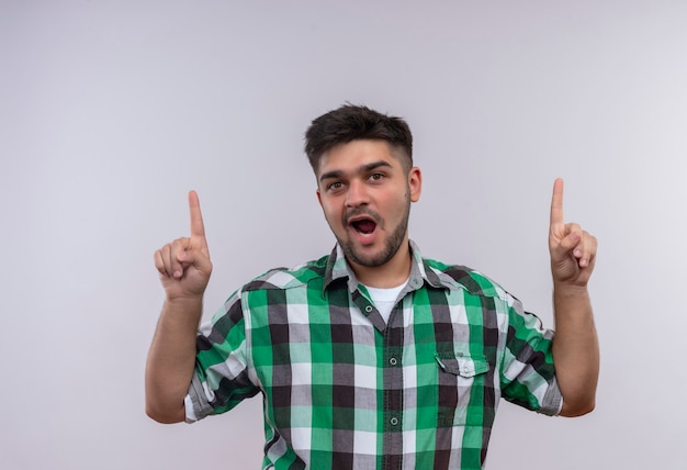 Young handsome guy wearing checkered shirt looking playfully and pointing forefingers up standing over white wall