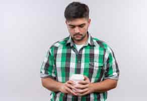 Foto gratuita giovane bel ragazzo che indossa la camicia a scacchi guardando tristemente verso il basso sulla sua tazza di caffè di plastica in piedi sopra il muro bianco