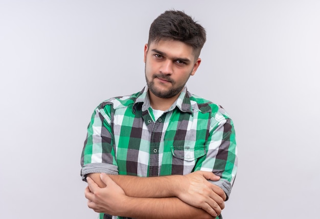 Young handsome guy wearing checkered shirt looking seriously standing over white wall
