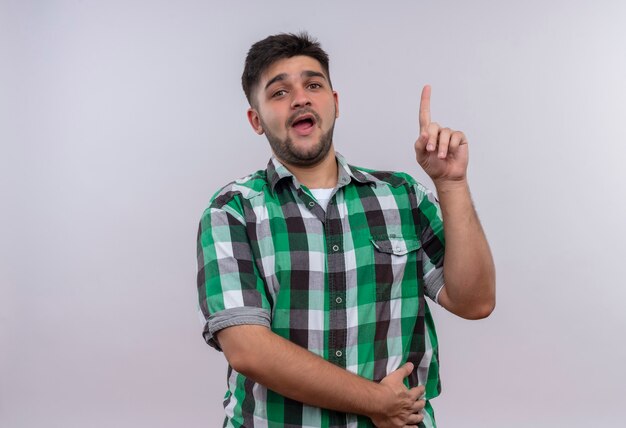 Young handsome guy wearing checkered shirt looking surprised pointing up with forefinger standing over white wall