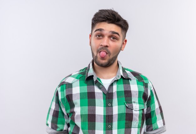 Young handsome guy wearing checkered shirt playfully looking showing tongue standing over white wall