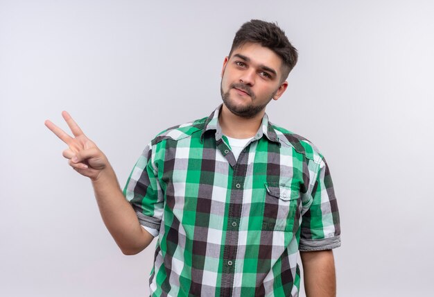 Young handsome guy wearing checkered shirt smiling doing peace sign with fingers standing over white wall