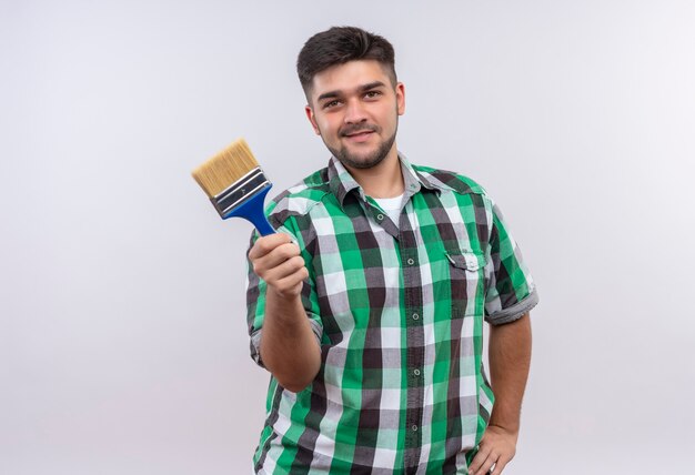 Young handsome guy wearing checkered shirt smiling holding paint brush standing over white wall