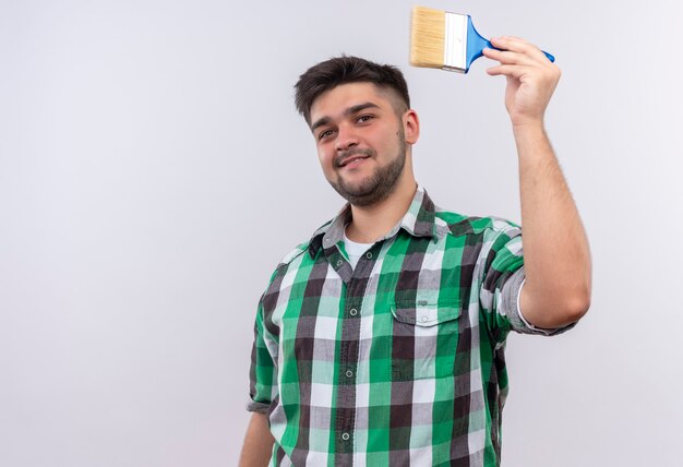 Young handsome guy wearing checkered shirt smiling painting with paint brush standing over white wall