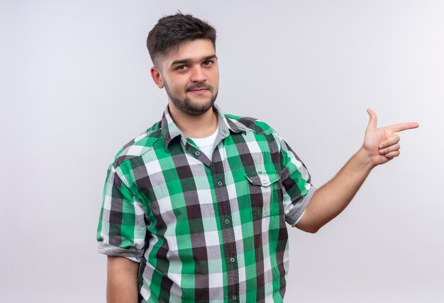 Young handsome guy wearing checkered shirt smiling pointing to the left standing over white wall