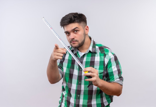 Foto gratuita giovane ragazzo bello che indossa il misuratore permanente della tenuta della camicia a scacchi che indica sopra il muro bianco