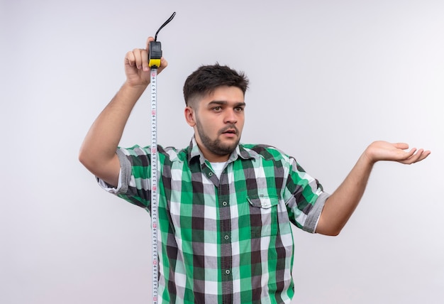 Free photo young handsome guy wearing checkered shirt undecided holding measurer looking besides doesn't know what to do standing over white wall