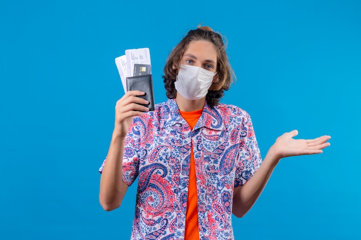 Free Photo young handsome guy wearing facial protective mask holding air tickets clueless and confused having no answer spreading hands standing