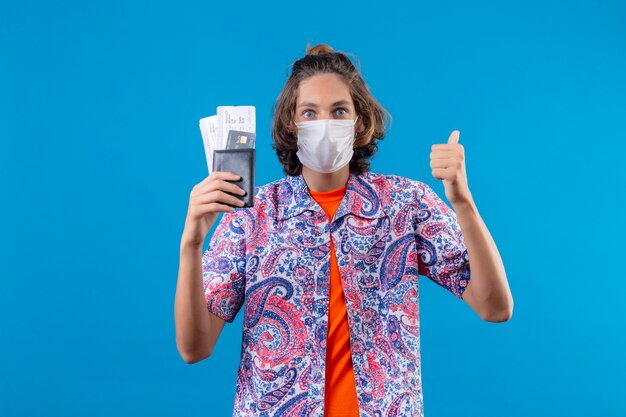 Young handsome guy wearing facial protective mask holding air tickets looking surprised showing thumbs up smiling cheerfully standing over blue background